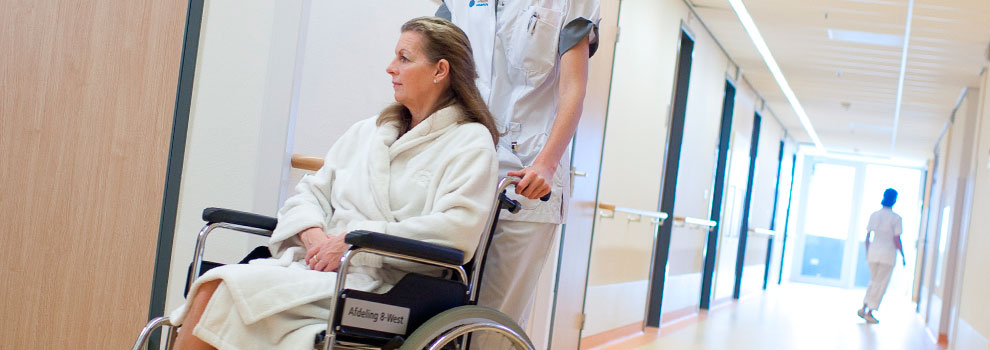 Patiënt in rolstoel, geduwd door verpleegkundige in de gang van het Catharina Ziekenhuis met op de achtergrond nog een verpleegkundige